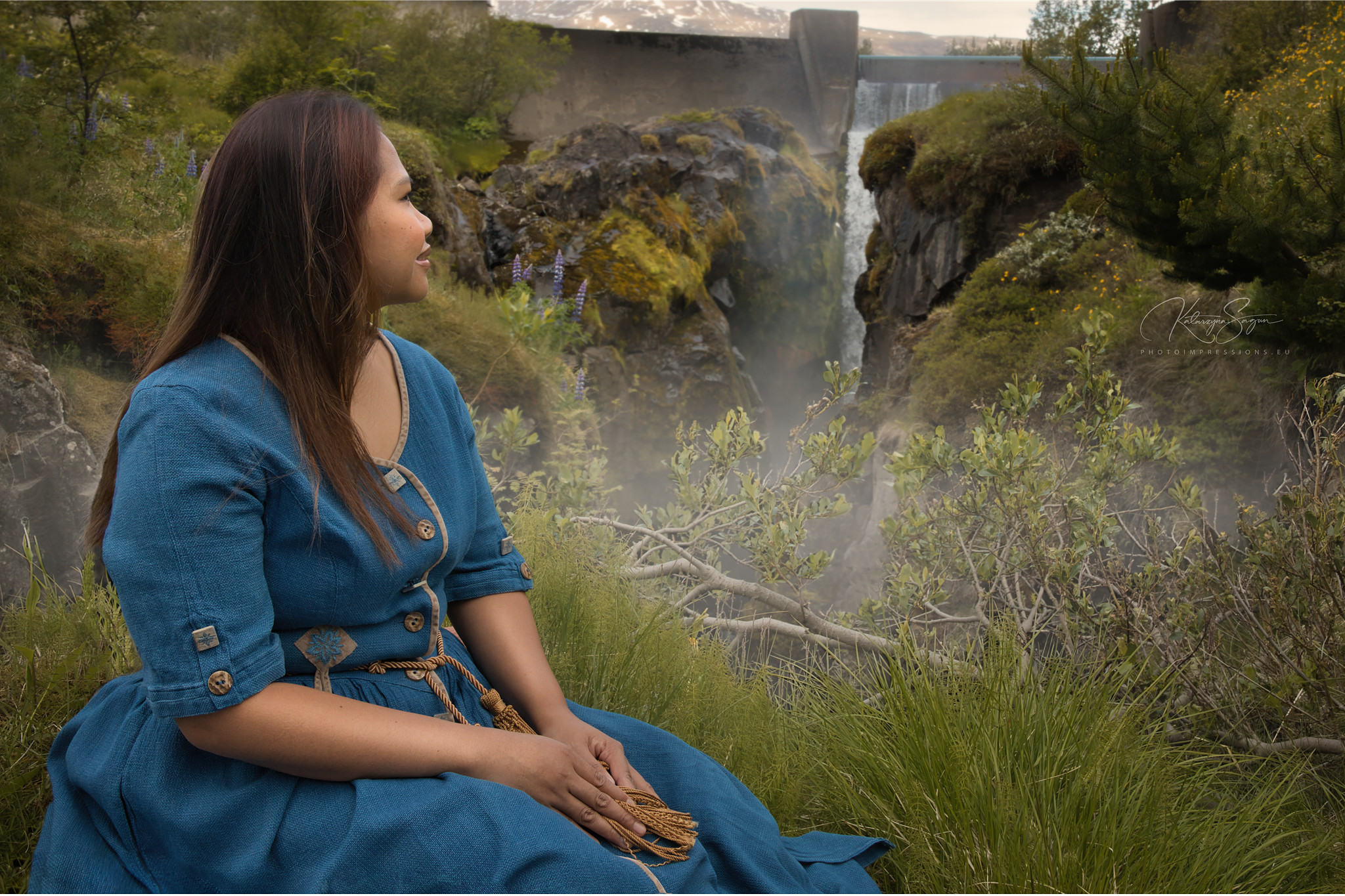 Woman in a long blue dress sitting by a stream, waterfall in Iceland.